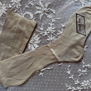Puede incluir: Una media de color beige, de estilo vintage, con una etiqueta que dice "Simili Soie". La media está sobre una superficie de encaje blanco con motivos florales.