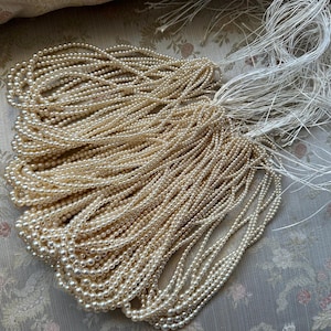 Puede incluir: Una colecci&oacute;n de collares de perlas blanquecinas con extremos de hilo blanco. Las perlas son peque&ntilde;as y redondas, y los collares est&aacute;n dispuestos en un mont&oacute;n suelto. El fondo es una tela con estampado floral.