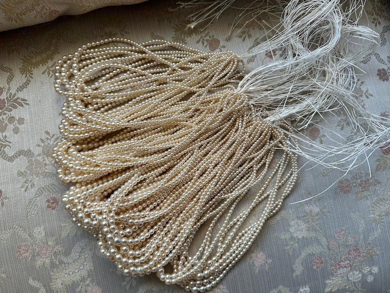 Puede incluir: Una colecci&oacute;n de collares de perlas blanquecinas con extremos de hilo blanco. Las perlas son peque&ntilde;as y redondas, y los collares est&aacute;n dispuestos en un mont&oacute;n suelto. El fondo es una tela con estampado floral.