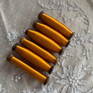 May include: Six spools of bright orange thread are arranged diagonally on a white lace background. The thread is tightly wound on wooden spools with dark brown ends. The lace has a floral pattern.