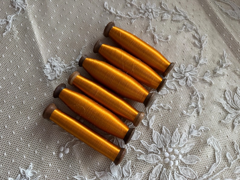 May include: Six spools of bright orange thread are arranged diagonally on a white lace background. The thread is tightly wound on wooden spools with dark brown ends. The lace has a floral pattern.