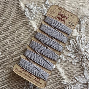 K&ouml;nnte beinhalten: Ein B&uuml;ndel grauer Stickgarnspulen mit Vintage-Etiketten. Die Etiketten tragen die Aufschrift "SOIE D'ALGER" und "MANUFACTURE DE SOIE A COUDRE & ABRODER". Der Faden ist auf kleinen Pappspulen aufgewickelt, vor einem Hintergrund aus zarter Spitze.