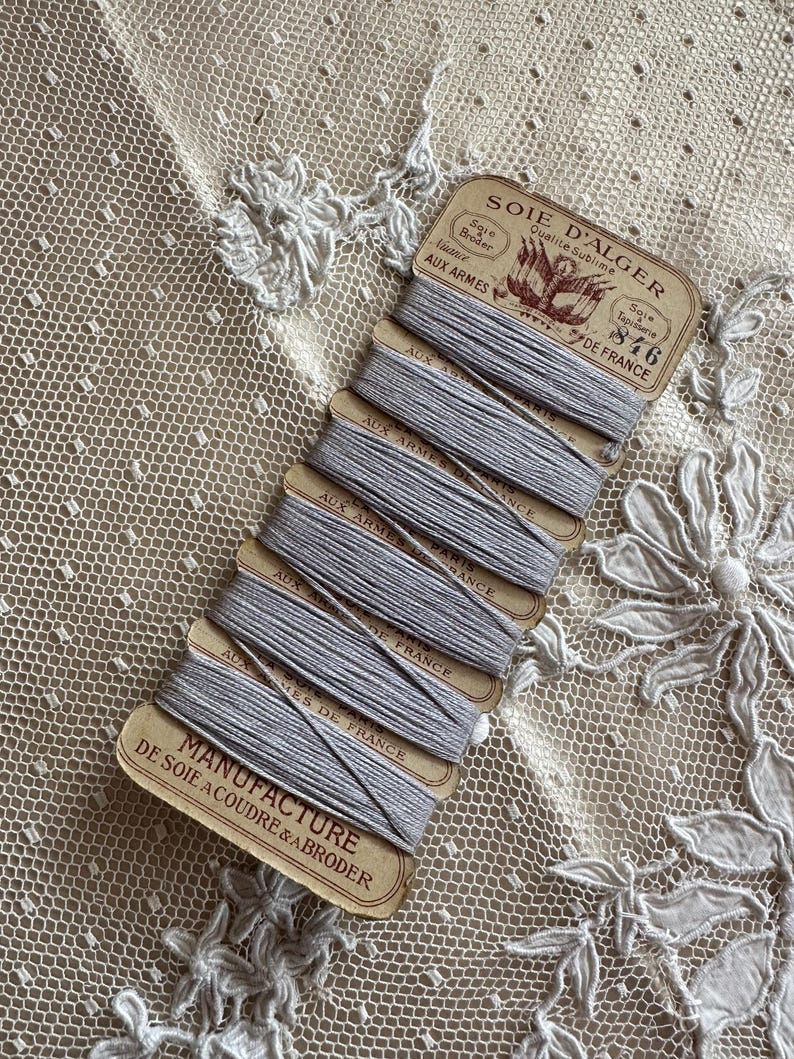 K&ouml;nnte beinhalten: Ein B&uuml;ndel grauer Stickgarnspulen mit Vintage-Etiketten. Die Etiketten tragen die Aufschrift "SOIE D'ALGER" und "MANUFACTURE DE SOIE A COUDRE & ABRODER". Der Faden ist auf kleinen Pappspulen aufgewickelt, vor einem Hintergrund aus zarter Spitze.