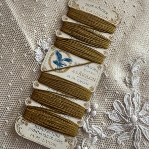 K&ouml;nnte beinhalten: Eine Sammlung von Vintage-Spulen aus olivgr&uuml;ner Seide. Jede Spule ist mit einem verzierten Text beschriftet, darunter "SOIE D'ALGER A L'AIGLON" und "MANUFACTURE LYONNAISE DE SOIE M.M. LYON". Die Spulen sind auf einem zarten, cremefarbenen Spitzengrund angeordnet.