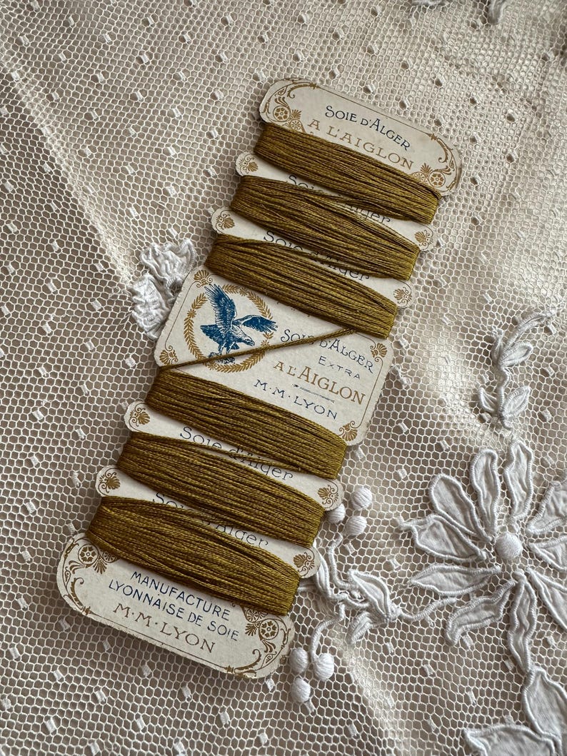 K&ouml;nnte beinhalten: Eine Sammlung von Vintage-Spulen aus olivgr&uuml;ner Seide. Jede Spule ist mit einem verzierten Text beschriftet, darunter "SOIE D'ALGER A L'AIGLON" und "MANUFACTURE LYONNAISE DE SOIE M.M. LYON". Die Spulen sind auf einem zarten, cremefarbenen Spitzengrund angeordnet.