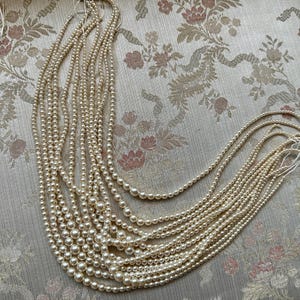 Puede incluir: Una colecci&oacute;n de collares de perlas de color crema de varias longitudes. Las perlas est&aacute;n ensartadas, creando un efecto en cascada. Los collares se exhiben sobre una tela con estampado floral.