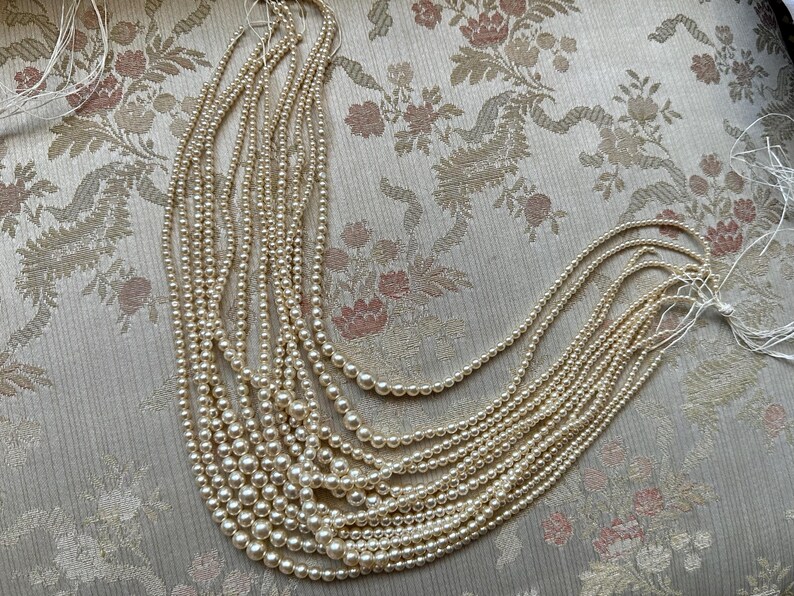 Puede incluir: Una colecci&oacute;n de collares de perlas de color crema de varias longitudes. Las perlas est&aacute;n ensartadas, creando un efecto en cascada. Los collares se exhiben sobre una tela con estampado floral.