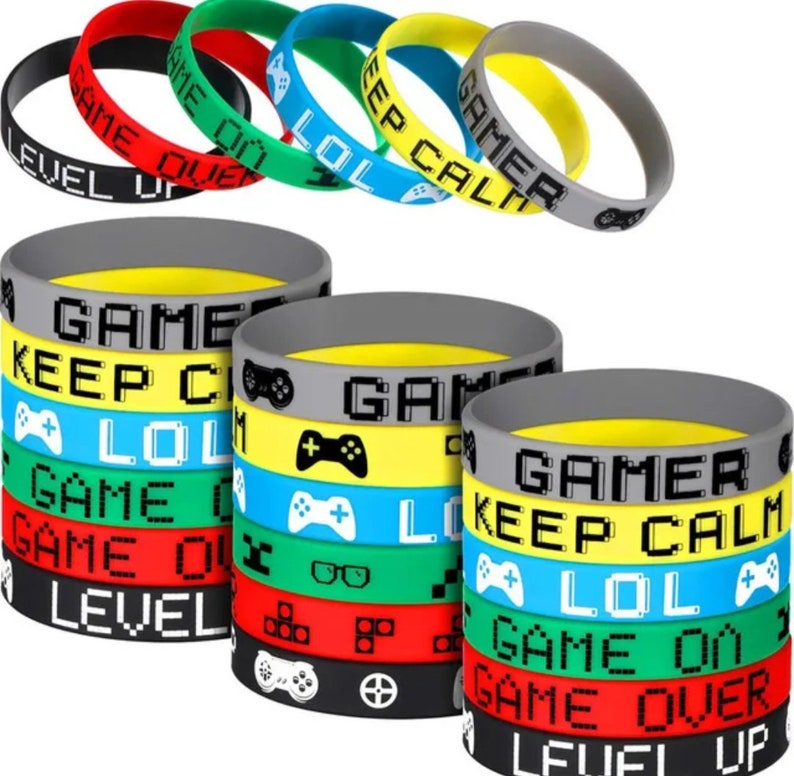 Może przedstawiać: Zestaw 10 kolorowych silikonowych bransoletek z motywami gier i napisami. Bransoletki zawierają takie frazy jak "Game Over", "Level Up", "Keep Calm", "Gamer" i "LOL".