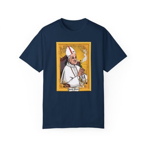 May include: A navy blue t-shirt featuring a graphic of a cartoon Pope smoking a joint and holding a marijuana leaf. The graphic is framed in gold and has the text "Dope Pope" at the bottom.