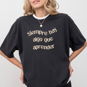 May include: A black t-shirt with the Spanish phrase "Siempre hay algo que aprender" (Always there is something to learn) printed in gold on the front.