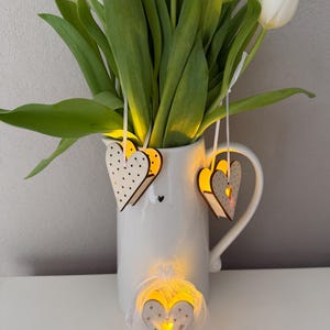 Könnte beinhalten: Eine weiße Keramikkanne mit einem Strauß weißer Tulpen. An der Kanne hängen drei hölzerne herzförmige Anhänger. Die Anhänger haben ein weißes Punkte-Muster und sind mit warmweißen LED-Lichtern beleuchtet. Einer der Anhänger liegt auf einer weißen Oberfläche und trägt den Text "Hello sunshine".