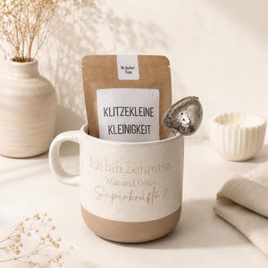 Könnte beinhalten: Ein zweifarbiger Keramikbecher mit gesprenkeltem cremefarbenem Oberteil und beigem Boden, bedruckt mit dem Text Ich bin Lehrerin. Was sind Deine Superkräfte? Ein brauner Papierbeutel mit der Aufschrift Kräuter Tee und Klitzekleine Kleinigkeit sowie ein herzförmiges Teesieb.