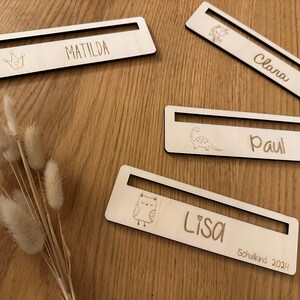 Könnte beinhalten: Vier Holznamensschilder mit einem Schlitz für ein Bild. Jedes Schild hat ein anderes Design und einen anderen Namen: Matilda, Clara, Paul und Lisa. Das Lisa-Schild hat auch den Text "Schulkind 2024".