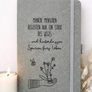 Könnte beinhalten: Ein graues Notizbuch mit vertikalem Gummiband und einer schwarzen Strichzeichnung einer Hand, die Blumen hält, sowie einer kleinen Biene. Auf dem Einband steht der Text: "MANCHE MENSCHEN BEGLEITEN NUR EIN STÜCK DES WEGES ... und hinterlassen Spuren fürs Leben."