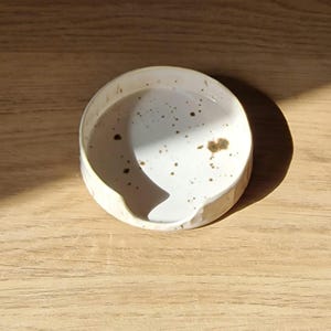 Pode incluir: Um prato de cerâmica redondo e raso com interior esmaltado em branco, apresentando manchas castanhas dispersas. O prato tem um rebordo liso e ligeiramente irregular com um pequeno entalhe curvo num dos lados, servindo de apoio para colher.