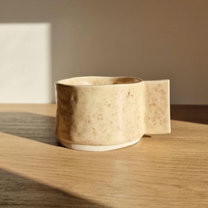 Op de afbeelding: Een beige keramische mok met een unieke, onregelmatige vorm en een handvat dat aan de zijkant uitsteekt. De mok heeft een gespikkelde, textuur.