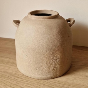 Pode incluir: Um vaso de cerâmica bege com uma superfície texturizada e duas pequenas alças. O vaso tem um corpo largo e arredondado e uma abertura estreita. O interior do vaso é escuro. O vaso está sobre uma superfície de madeira.