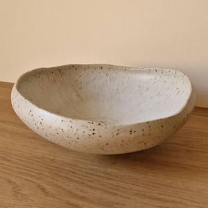Pode incluir: Tigela de cerâmica branca suja com manchas escuras. Com formato irregular e orgânico, esta tigela rasa é ideal para servir alimentos ou como elemento decorativo. Feito à mão.