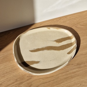 Pode incluir: Um prato de servir oval de cerâmica branca com veios marrons. O prato tem uma superfície ligeiramente texturizada.