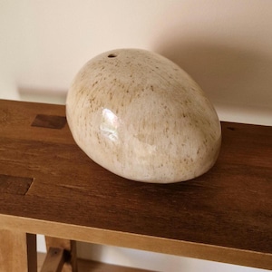 Pode incluir: Uma pedra decorativa lisa, de forma oval, com um acabamento bege salpicado. A pedra tem um pequeno orifício na parte superior e está sobre uma superfície de madeira. A superfície polida da pedra reflete a luz, adicionando um brilho subtil. Cerca de 20 cm de comprimento.