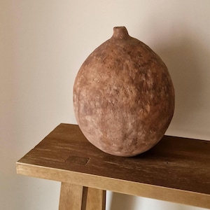 Puede incluir: Un jarrón decorativo marrón grande y texturizado, con un cuerpo redondeado y un cuello pequeño y cilíndrico. El jarrón se exhibe sobre un banco de madera. El jarrón tiene una estética rústica y terrosa.