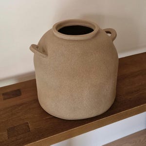 Pode incluir: Um vaso de cerâmica bege, redondo, com uma abertura larga e duas pequenas alças. O vaso tem uma superfície texturizada e está sobre uma prateleira de madeira. O interior do vaso é escuro.
