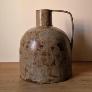 Peut inclure: Une cruche en grès avec un corps arrondi, un col cylindrique et une anse. La cruche a un aspect moucheté et marbré dans des tons bruns et gris. La poterie faite à la main est probablement à usage décoratif.
