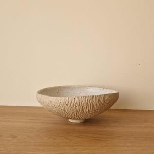Può includere: Una ciotola in ceramica bianco sporco con superficie strutturata e interno maculato. La ciotola ha una forma ovale e poggia su una piccola base rotonda. L'esterno presenta un motivo verticale a coste.