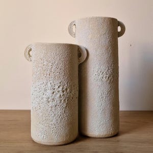 Peut inclure: Deux vases en céramique blanc cassé avec une surface texturée et de petites anses. Les vases sont cylindriques, l'un étant plus grand que l'autre. La surface a un aspect moucheté, ajoutant un charme rustique. Ces vases décoratifs conviennent à la décoration intérieure.