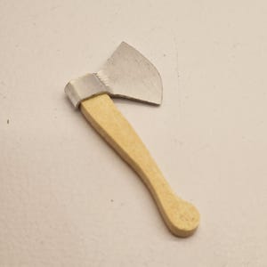 Pode incluir: Um machado em miniatura com uma lâmina prateada e um cabo de madeira castanho claro. O cabo é curvo. A cabeça do machado tem aproximadamente 2,5 cm de largura.