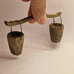 Könnte beinhalten: Eine Miniatur-Holzwaage mit zwei kleinen Eimern, die an Draht aufgehängt sind. Die Waage wird von einer Hand vor einem neutralen Hintergrund gehalten. Die Eimer und der Waagebalken sind aus geschnitztem Holz.