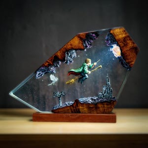 May include: A decorative resin sculpture featuring a boy on a broomstick, a castle, and a bird against a starry background. The sculpture is encased in a clear, angled frame with a wooden base and accents. The boy wears a green cloak.