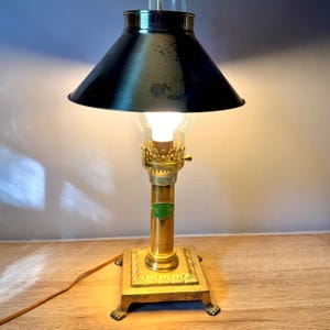 May include: A brass table lamp featuring a conical dark metal shade, a clear glass chimney, and a central column with a green oval label. The square base has decorative edges and four claw-style feet, with a thin brown power cord extending outward.