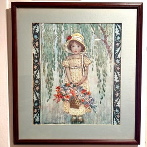 May include: A framed embroidery art piece depicting a child in a yellow bonnet and dress holding a basket of colorful flowers. The scene is set against a backdrop of green willow branches, framed by a dark wood border and a light blue matting.