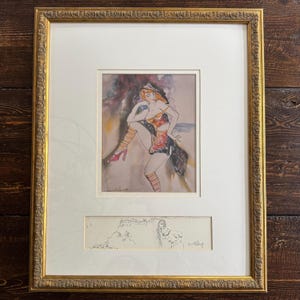 May include: Framed artwork featuring a watercolor and ink illustration of a dancer in a colorful costume. The artwork is in a gold frame and includes a smaller sketch below the main image. The dancer is wearing a hat, heels, and striped stockings.