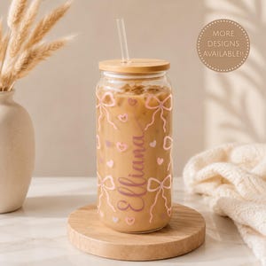 Pode incluir: Um copo transparente com tampa de bambu e palha transparente, cheio de café gelado. O copo apresenta um padrão gráfico rosa de laços e pequenos corações, com o nome Elliana escrito verticalmente numa fonte cursiva de cor rosa.