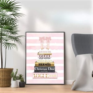 May include: A framed print featuring a stack of fashion books with designer names like Gucci, Hermes, and Christian Dior. The artwork includes a pair of high-heeled boots and a pink and white striped background. The Vogue 100 Years logo is visible.