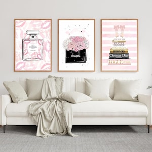May include: Three framed art prints with fashion illustrations. One depicts a perfume bottle, another a bouquet in a bag, and the third a stack of designer books and shoes. The prints feature pink and black accents.