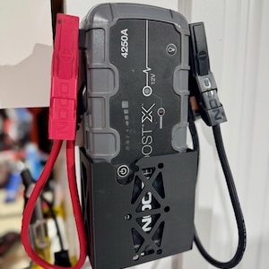 Puede incluir: Un arrancador de batería negro y gris con un soporte de montaje negro. El arrancador de batería tiene una pantalla digital y el texto "BOOST X" y "4250A" impreso en él. El arrancador de batería tiene cables de arranque rojo y negro conectados a él.