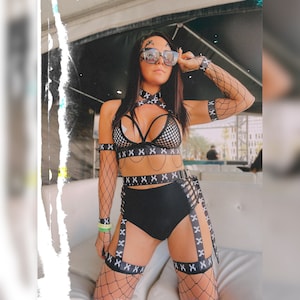 Puede incluir: Un conjunto de lencería negro y blanco con un top de malla y una parte de abajo de cintura alta. El conjunto presenta un diseño de correas cruzadas y un cinturón negro con una hebilla plateada. La modelo lleva medias de rejilla negras y gafas de sol.