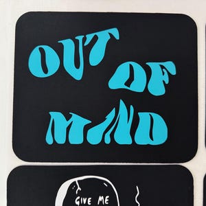 以下が含まれることがあります： 黒いステッカーで、ターコイズ色のスタイリッシュな文字で「OUT OF MIND」と書かれています。ステッカーは角が丸い長方形です。テキストは太字で波状のフォントで、レトロな美学を作り出しています。ステッカーはおそらく装飾目的です。