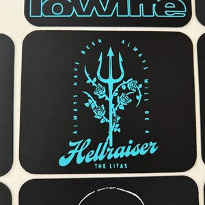 May include: Black square sticker with a teal trident design, roses, and the words "ALWAYS HAVE BEEN ALWAYS WILL BE A" around the trident. Below is the word "Hellraiser" and "THE LITAS" in teal.