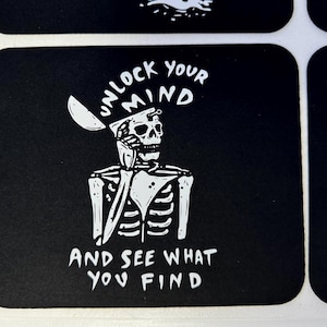 Może przedstawiać: Czarno-biała naklejka z motywem szkieletu. Tekst brzmi "UNLOCK YOUR MIND" nad głową szkieletu i "AND SEE WHAT YOU FIND" poniżej. Naklejka ma zaokrąglone rogi.