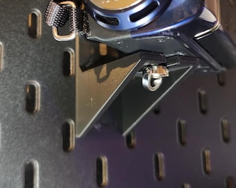 May include: A black plastic wall-mounted bracket attached to a dark gray pegboard, supporting a blue Sony camcorder. The bracket features a triangular support structure and a silver metal ring attachment point positioned directly beneath the camera.