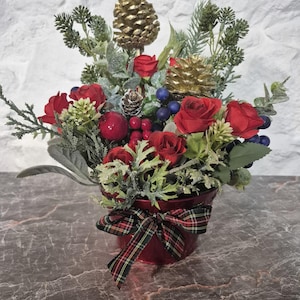 May include: A floral arrangement in a red metal pot featuring red roses, gold-painted pinecones, green foliage, and small blue berries. The pot is decorated with a red, green, and gold plaid ribbon tied in a bow at the front of the container.