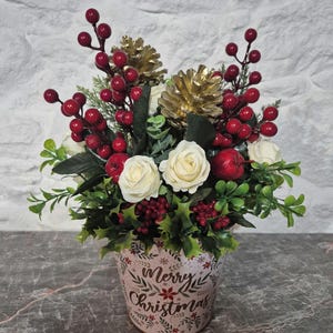 May include: A floral arrangement in a light-coloured bucket with the text Merry Christmas. The arrangement features white roses, red berries, green foliage, gold-painted pinecones, and small red apples, creating a festive holiday display.