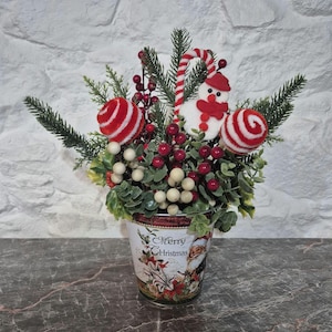 May include: A decorative arrangement in a metal bucket featuring the text Merry Christmas, filled with green pine branches, red and white striped spheres, white berries, red berries, and a white snowman ornament with a red hat and scarf.