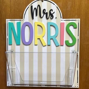 以下が含まれることがあります： 「Mrs. Norris」の文字が黒い筆記体で書かれ、その下にカラフルなブロック体で「NORRIS」と書かれた木製のサイン。サインには、小物を入れるための透明なプラスチックポケットが付いています。背景はベージュと白の縦縞模様です。