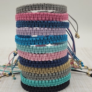 May include: A stack of 12 colorful friendship bracelets made with woven cord. The bracelets are in various shades of blue, pink, gray, and teal. Each bracelet has a gold bead at the end of the cord.
