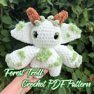 Może przedstawiać: Biały szydełkowy Forest Troll z zielonymi akcentami i brązowymi rogami. Pluszowa zabawka ma duże czarne oczy i zielone akcenty. Tekst "Forest Troll Crochet PDF Pattern" jest wyświetlany w kolorze zielono-białym.